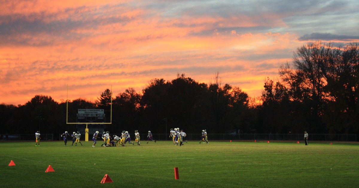 A vivid orange sunset over a football field during a game, players in silhouette